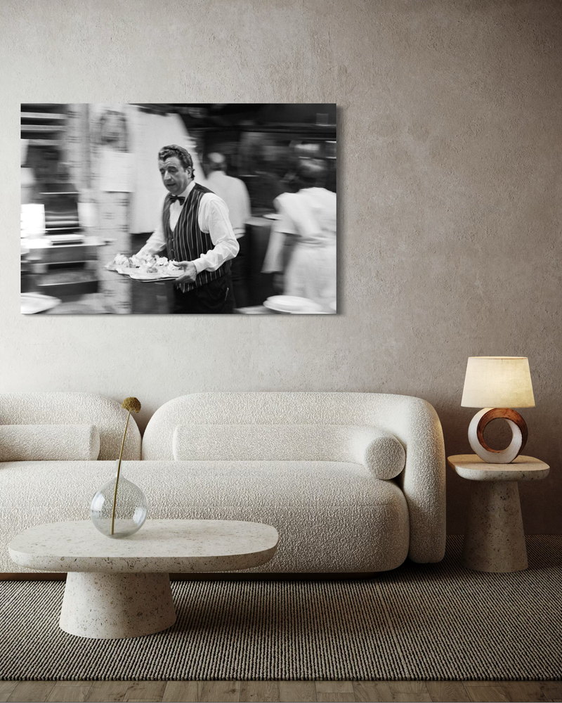 Zwart-wit fotografie op canvas van een ober in beweging die een dienblad door een restaurant draagt, met vervaagde achtergrond en gevoel van tempo.